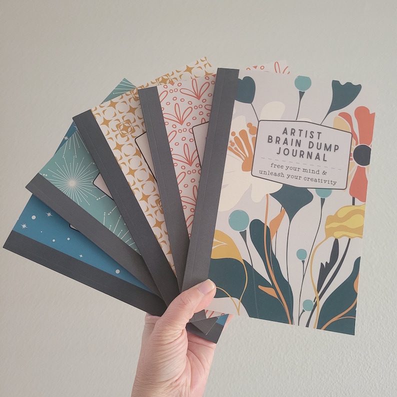 May include: A stack of five journals with different floral and star patterns. The top journal has a floral design and the text "Artist Brain Dump Journal" with the tagline "free your mind & unleash your creativity".