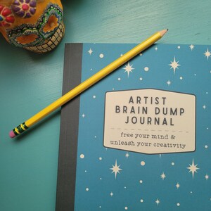 May include: A blue journal with white stars and the text "Artist Brain Dump Journal" and "free your mind & unleash your creativity". A yellow pencil with green and black stripes is lying on top of the journal.