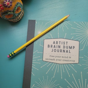 May include: A teal blue journal with a black spine and a yellow pencil on top. The journal cover has a repeating pattern of white starbursts and the text "Artist Brain Dump Journal" with the tagline "free your mind & unleash your creativity".