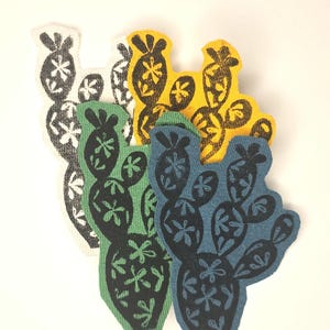 May include: A collection of cactus-shaped fabric patches in various colors. The patches feature a black floral design on backgrounds of white, green, yellow, and blue. The patches are arranged in a layered pattern.