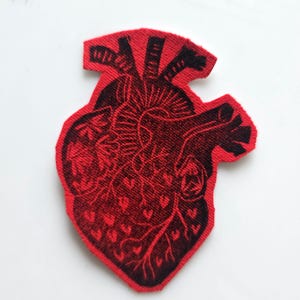 May include: A red and black embroidered patch in the shape of a human heart. The patch features a detailed anatomical design of the heart's chambers, vessels, and veins. The heart's outline and internal details are in black.