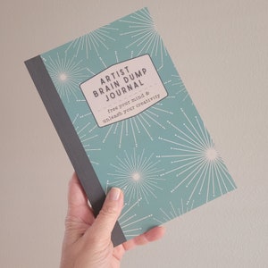 May include: A teal blue journal with a white and gray starburst pattern. The cover has a white rectangle with the text "Artist Brain Dump Journal" and the text "free your mind & unleash your creativity".