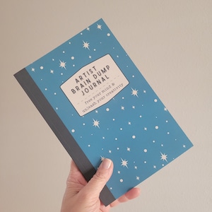 May include: A blue journal with white stars printed on the cover. The journal is titled "Artist Brain Dump Journal" and has the text "free your mind & unleash your creativity" printed on the cover.