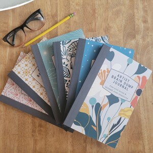 May include: A stack of six colorful journals with floral and geometric designs. The top journal has the text "Artist Brain Dump Journal - Free your mind & unleash your creativity".