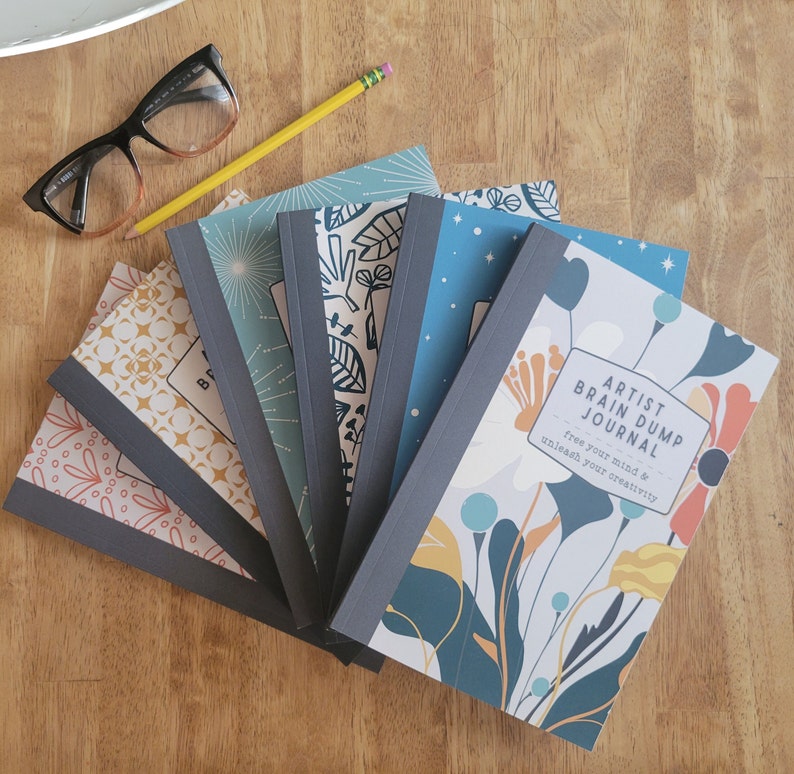 May include: A stack of six colorful journals with floral and geometric designs. The top journal has the text "Artist Brain Dump Journal - Free your mind & unleash your creativity".