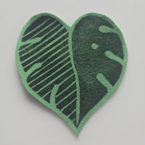 May include: A heart-shaped patch featuring a green leaf design. One half of the leaf has black diagonal stripes, while the other half has a monstera leaf pattern. The patch has a green border.