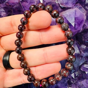 May include: A beaded bracelet featuring dark red garnet gemstones. The round beads are strung together, creating a bracelet. The bracelet is held against a background of purple crystals.
