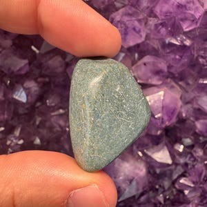 May include: A smooth, rounded, teal-colored stone with subtle marbling is held between two fingers. The background is a cluster of purple amethyst crystals. The stone appears to be a tumbled gemstone, possibly for use in jewelry or crystal healing.