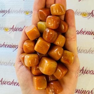 May include: A hand holding a pile of tumbled orange carnelian stones. The stones are smooth and polished, and they have a warm, inviting color.