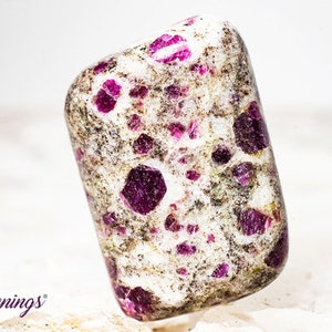 May include: A white tumbled stone with a high concentration of small, deep pink ruby inclusions. The stone has a speckled appearance.