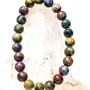 May include: A bracelet made of round, multi-colored gemstone beads. The beads are a mix of green, brown, purple, and blue. The bracelet is displayed on a white crystal.