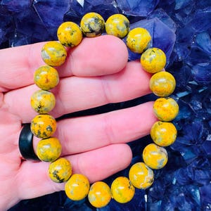 Könnte beinhalten: Ein gelbes und schwarzes Bumblebee-Jaspis-Armband mit Perlen. Das Armband besteht aus runden Perlen und wird auf einer Hand gezeigt.