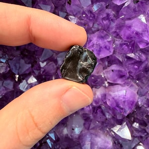 Pode incluir: Uma pequena pedra escura de forma irregular segurada entre os dedos, contra um fundo de vibrantes cristais de ametista roxa. A pedra parece ser um esp&eacute;cime mineral polido.