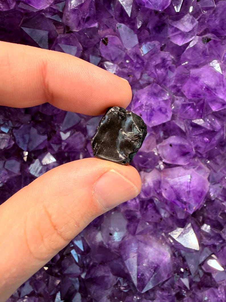 Pode incluir: Uma pequena pedra escura de forma irregular segurada entre os dedos, contra um fundo de vibrantes cristais de ametista roxa. A pedra parece ser um esp&eacute;cime mineral polido.