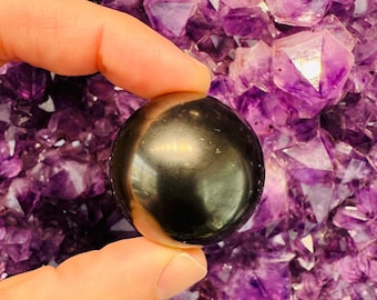 May include: A smooth, black obsidian sphere, also known as a crystal ball, is held in a hand against a background of purple amethyst crystals.