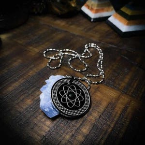 May include: A silver chain necklace with a black pendant featuring an atom symbol. The pendant is layered over a light blue crystal.