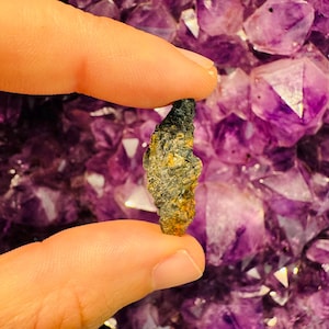 Puede incluir: Una pequeña roca de color gris oscuro y marrón con una forma rugosa e irregular. La roca está siendo sostenida por un dedo contra un fondo de cristales de amatista púrpura.