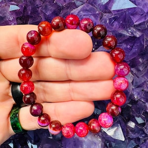 May include: A beaded bracelet featuring a variety of red and pink gemstones. The round beads display a range of shades, from deep burgundy to vibrant pink. The bracelet is held against a purple background.