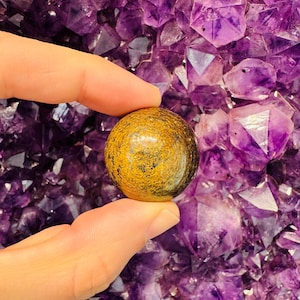 May include: A golden tiger's eye sphere with brown and black stripes, resting on a hand against a background of purple amethyst crystals.