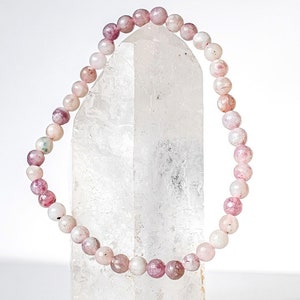 May include: A pink and white gemstone bracelet with small round beads. The bracelet is displayed on a clear quartz crystal.