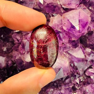 Puede incluir: Una piedra preciosa ovalada de color rojo oscuro con una superficie brillante y pulida. La piedra preciosa se sostiene en una mano sobre un fondo de cristales de amatista púrpura.
