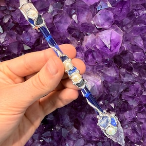 May include: A handcrafted wand with a clear crystal tip and a blue and silver handle. The wand is adorned with pearls and other crystals. The background is a cluster of purple amethyst crystals.
