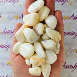 May include: A handful of polished, creamy white stones with a smooth, rounded shape. Some stones have subtle tan or brown markings. The stones are held in an open hand, with a soft, natural light illuminating them.