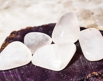 May include: Five smooth, white, polished stones, likely selenite, arranged on a dark, textured surface.