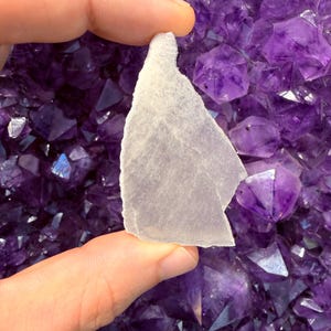 May include: A hand holds a translucent, light-colored, irregularly shaped crystal fragment against a backdrop of deep purple amethyst crystals. The amethyst crystals have sharp, geometric facets.