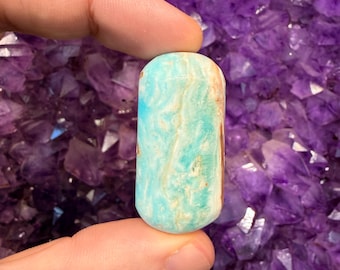 May include: A small, cylindrical gemstone with a rounded top. The stone features a mix of light blue, turquoise, and cream colors. The gemstone is held between two fingers against a background of purple crystals.