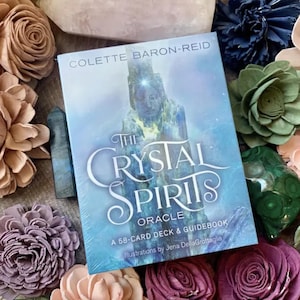 以下が含まれることがあります： 「The Crystal Spirits Oracle」というタイトルの青と白のオラクルデッキボックス。テキストは「A 58-card deck & guidebook. Illustrations by Jena Della Grottaglia.」