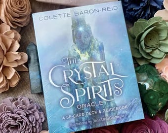 May include: A blue and white oracle deck box with the title "The Crystal Spirits Oracle" and the text "A 58-card deck & guidebook. Illustrations by Jena Della Grottaglia."