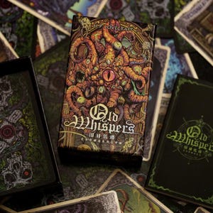 May include: A box of Old Whispers tarot cards featuring a dark green and black design with a multi-eyed creature on the front. The box is open and shows the cards inside, which are also dark green and black with a similar design.