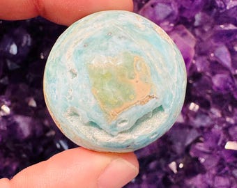 May include: A round, light blue stone with a central tan and green pattern. The stone has a textured surface and is held between two fingers against a background of purple crystals. The stone appears to be a decorative item or a natural specimen.