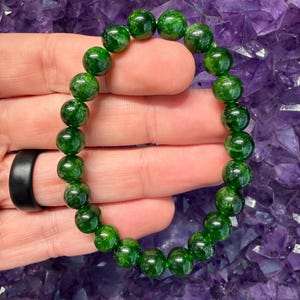 May include: A circular beaded bracelet composed of polished, translucent deep green gemstone spheres. The beads are uniform in size and strung together on an elastic cord, creating a continuous loop of vibrant green stones with natural internal inclusions.