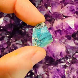 Może przedstawiać: Mały, turkusowy kryształ o szorstkiej, naturalnej fakturze. Kryształ jest trzymany przez palec na tle fioletowych kryształów ametystu.
