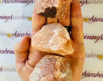 May include: Three rough, natural, orange and white crystal stones. The stones are in the palm of a hand.