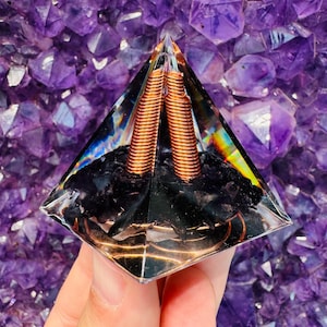 May include: A clear glass pyramid with a black stone base and two copper coils inside. The pyramid is sitting on a bed of purple amethyst crystals.