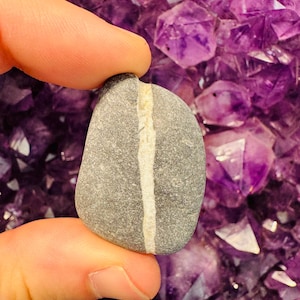 May include: A smooth, gray river rock with a white stripe running down the center. The rock is being held by a hand against a background of purple amethyst crystals.