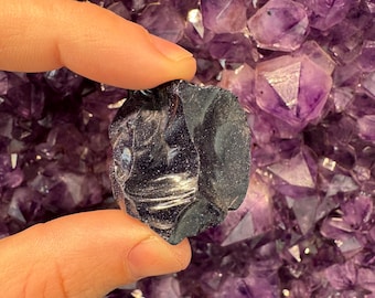 Puede incluir: Un cristal gris oscuro, de textura rugosa, con un brillo metálico. El cristal está siendo sostenido por una mano contra un fondo de cristales de amatista púrpura.