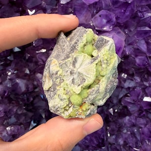 Pode incluir: Um espécime mineral bruto apresentando uma matriz cinza-escura com aglomerados de formações esféricas verde-pálidas e estruturas cristalinas radiantes em forma de leque. A superfície exibe uma mistura de texturas, incluindo nódulos lisos e arredondados.