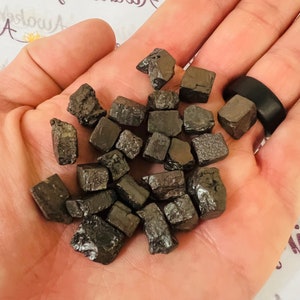 Puede incluir: Una colección de pequeños cristales de color gris oscuro en forma de cubo. Los cristales son ásperos y tienen un brillo metálico.