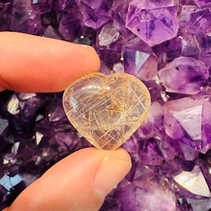 May include: A heart-shaped clear quartz crystal with gold inclusions. The crystal is held in a hand against a background of amethyst crystals.