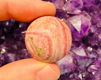May include: A small, spherical gemstone with a pink and white striped pattern, held between fingers. The stone has a polished surface and a few green spots. The background is a cluster of purple amethyst crystals.