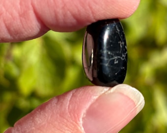 May include: A smooth, black, oval-shaped stone with a slight sheen. The stone is being held between two fingers.