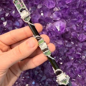 May include: A handcrafted wand featuring a black, polished central shaft with silver-toned metal accents. The wand is adorned with green, white, and red-speckled gemstones. The wand's tip is a dark, pointed crystal. The background is a cluster of purple amethyst crystals.