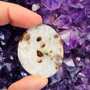 May include: A white, oval-shaped, polished stone with brown inclusions. The stone is being held by a hand against a background of purple amethyst crystals.