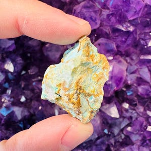 May include: A small, rough, turquoise-colored rock with yellow and brown markings. The rock is being held by a hand against a background of purple amethyst crystals.