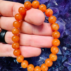 May include: A bracelet made of orange gemstone beads. The beads are round and have a polished, glossy finish. The bracelet is held against a background of purple crystals, creating a contrast of colors and textures.