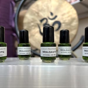 May include: Five small glass bottles with black caps, filled with a green liquid. Each bottle has a white label with the word "MOLDAVITE" and the text "EXTERNAL USE ONLY". The bottles are lined up on a white surface.